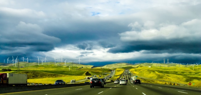 Eine dreispurige Autobahn mit Verkehr in beide Richtungen. Neben der Autobahn stehen mehrere Windräder zur Stromgewinnung.