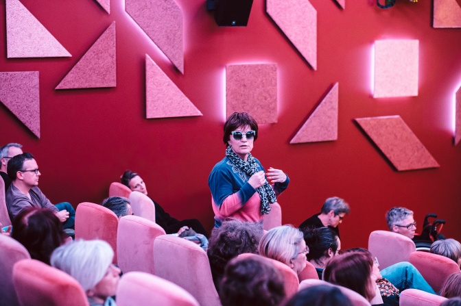 Eine blinde Frau mit dunkler Sonnenbrille geht an Sitzreihen eines Kinos vorbei.