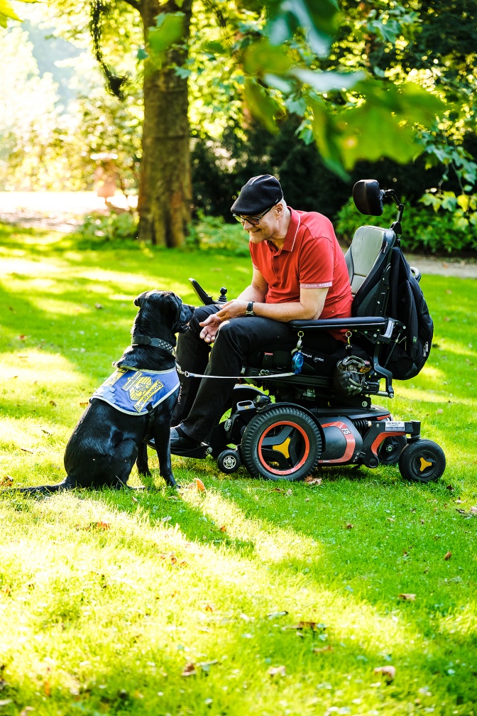 Ein Mann in einem elektrischen Rollstuhl auf einer Wiese, zusammen mit einem schwarzen Assistenzhund, der vor ihm im Gras sitzt.