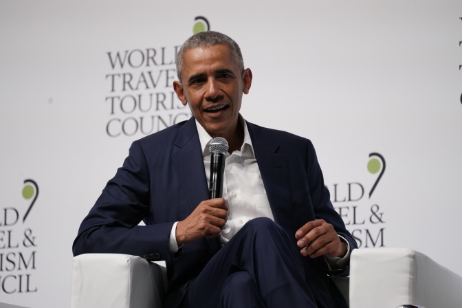 Barack Obama in Anzug und Krawatte. Er sitzt vor einer Logowand und spricht in ein Mikrophon.