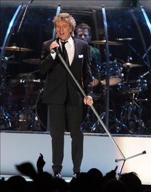 Rod Stewart steht auf der Bühne und singt in ein Standmikro. Er trägt Anzug und Krawatte.