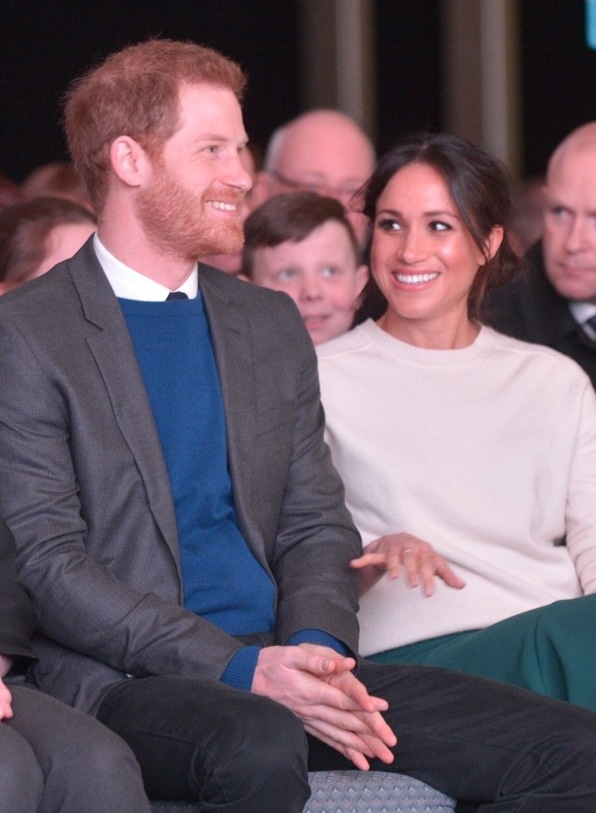 Prinz Harry und Herzogin Meghan sitzen nebeneinander, hinter ihnen sitzen andere Menschen. Beide lächeln.