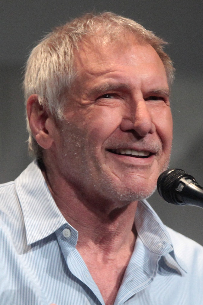 Harrison Ford in einem locker geknöpften Hemd und mit kurzen, grauen Haaren. Er hat einen Dreitagebart und sitzt vor einem Mikrophon.