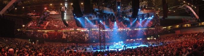 Ein Panorama-Foto der Band in einem sehr großen Publikum und mit fsrbiger Beleuchtung