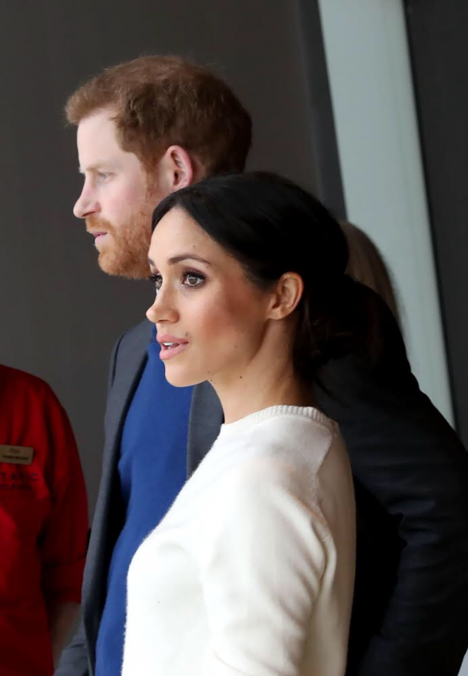 Prinz Harry und Herzogin Meghan. stehen hintereinander. Meghan trägt einen weißen Strickpullover und Harry ein blaues Sakko.