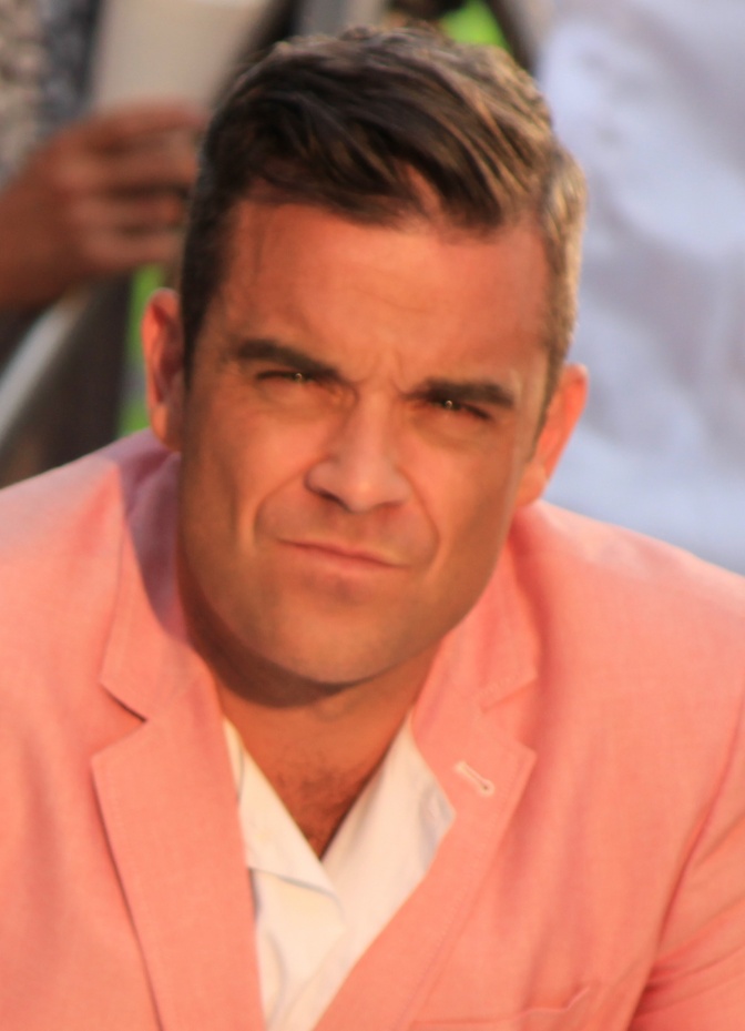 Robbie Williams in einem lachsfarbenen Sakko. Er guckt nachdenklich.