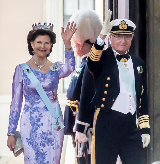 Die Königin trägt ein festliches, fliederfarbenes Kleid und der König eine Uniform. Beide winken und tragen mehrere Orden und Auszeichnungen.