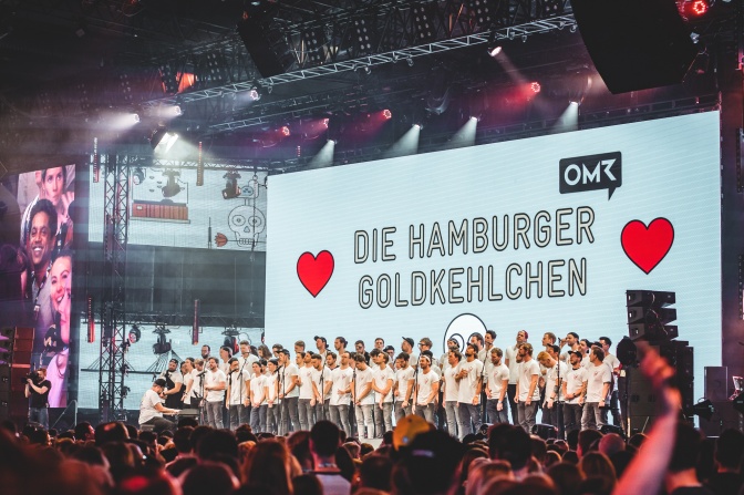70 Männer in einheitlichen T-Shirts stehen auf der Bühne vor einem großen Banner mit dem Text Die Hamburger Goldkehlchen