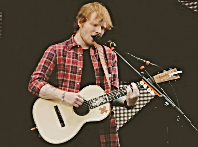 Ed Sheeran trägt ein Holzfällerhemd, hat rote Haare, spielt eine akustische Gitarre und singt dazu.