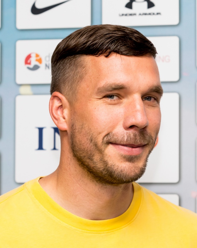 Lukas Podolski mit Bart und braunen, an den Seiten ausrasierten Haaren. Er trägt ein gelbes T-Shirt und steht vor einer Logowand.