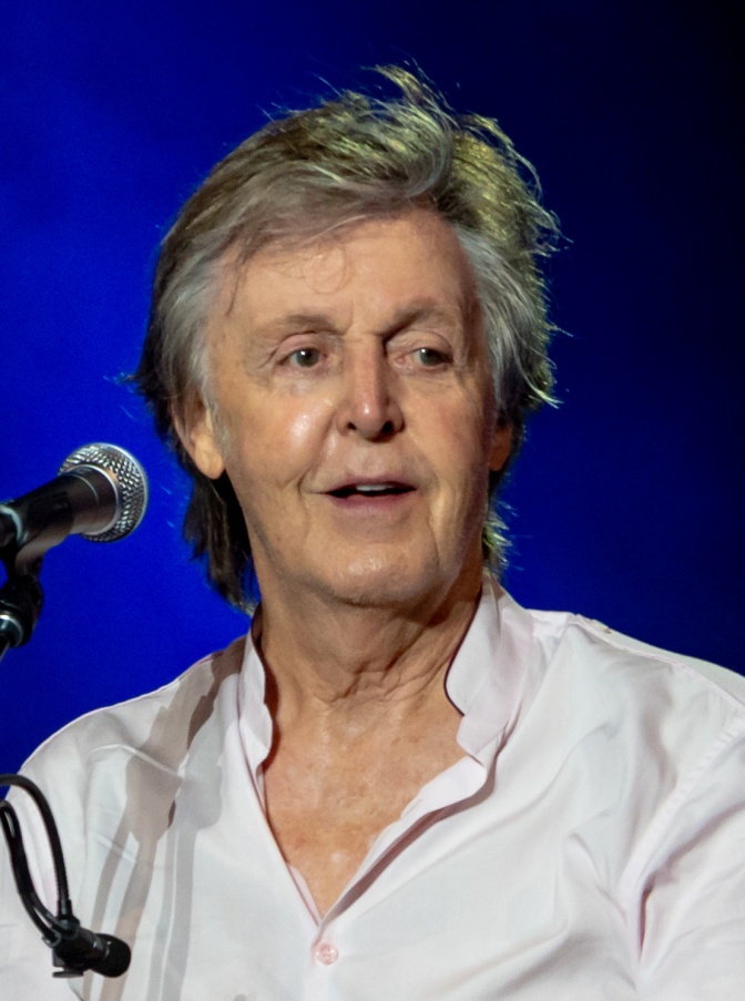 Paul McCartney mit grauen Haaren und in weißem Hemd vor einem Mikrophon.