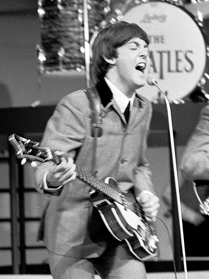 Ein schwarz-weiß-Foto von Paul McCartney beim Passspielen und Singen. Er  steht auf der Bühne. Im Hintergrund sieht man eine Base Drum mit dem Schriftzug der Beatles.