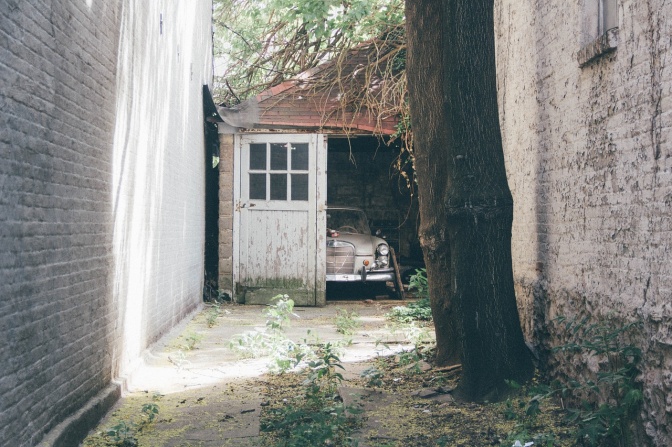Ein Oldtimer steht in einer Garage. Das Tor ist zur Hälfte geschlossen. Im Vordergrund steht ein Baum.