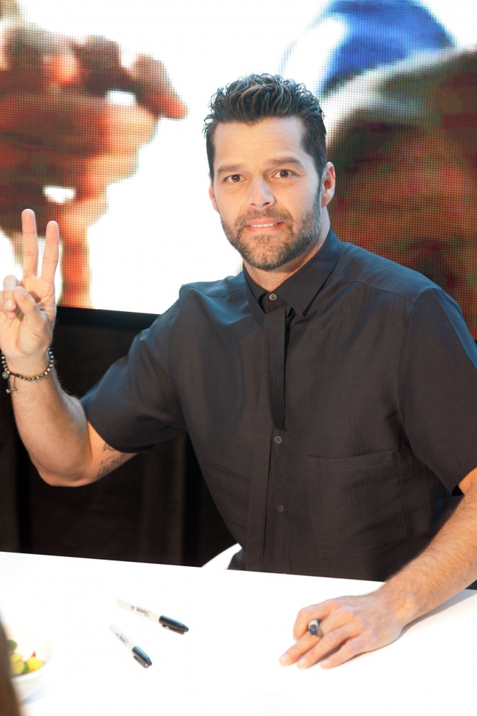 Ricky Martin sitzt an einem Tisch und hebt  die Hand zum Victory-Zeichen.
