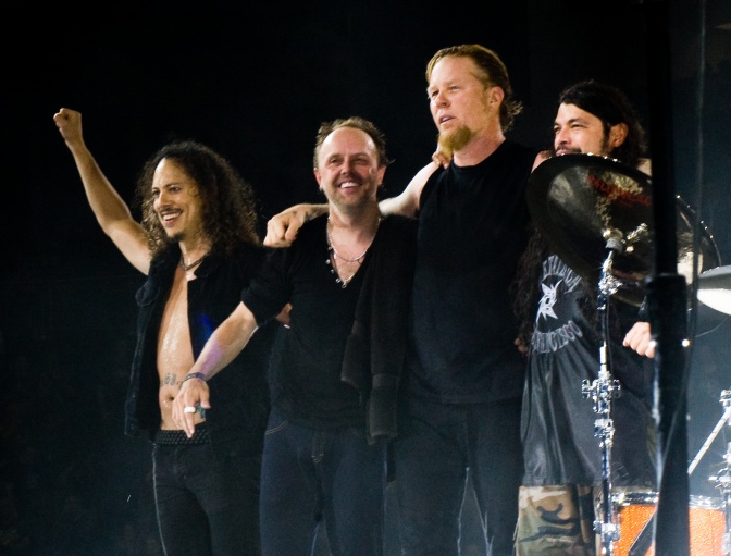 Die Mitglieder der Band Metallica auf der Bühne. Ein Bandenmitglied reckt den Arm in die Luft.