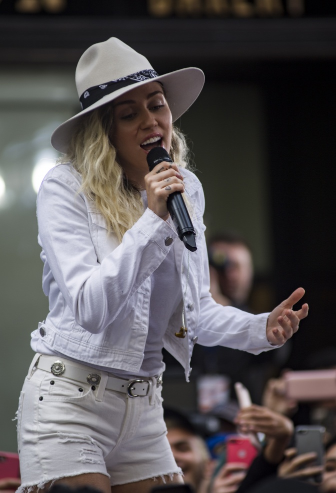 Miley Cyrus in weißer Shorts und mit weißem Cowboyhut auf der Bühne. Sie singt in ein Mikrophon.