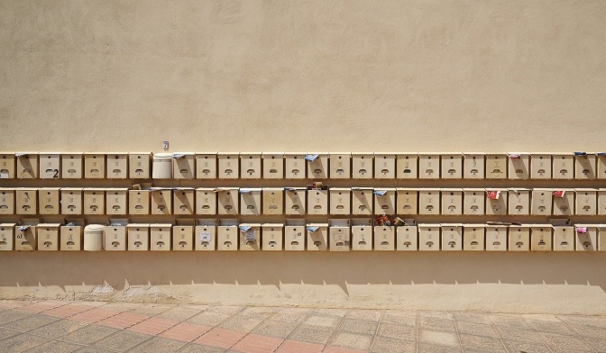 Sehr viele Briefkästen, in 3 Reihen untereinander an einer Wand angebracht. Aus manchen Briefkästen ragt Post heraus.