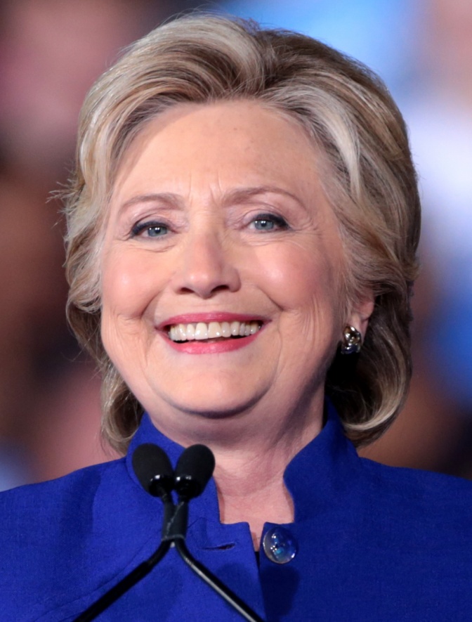 Hillary Clinton lächelt breit in die Kamera. Sie hat blonde Haare mit grauen Strähnen, zu einer Kurzhaarfrisur frisiert.
