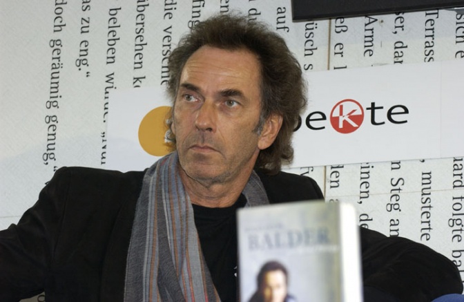 Hugo Egon Balder mit gelockten, dunklen Haaren. Er sitzt vor einer Logowand, vor ihm steht  aufgestellt eines seiner Bücher.