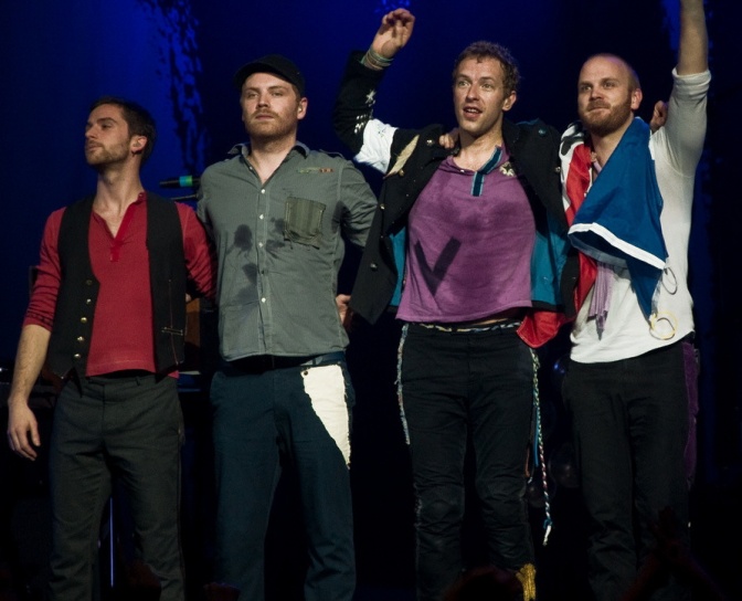 Die Mitglieder der Band Coldplay stehen auf der Bühne nebeneinander. Ein Bandenmitglied winkt ins Publikum.
