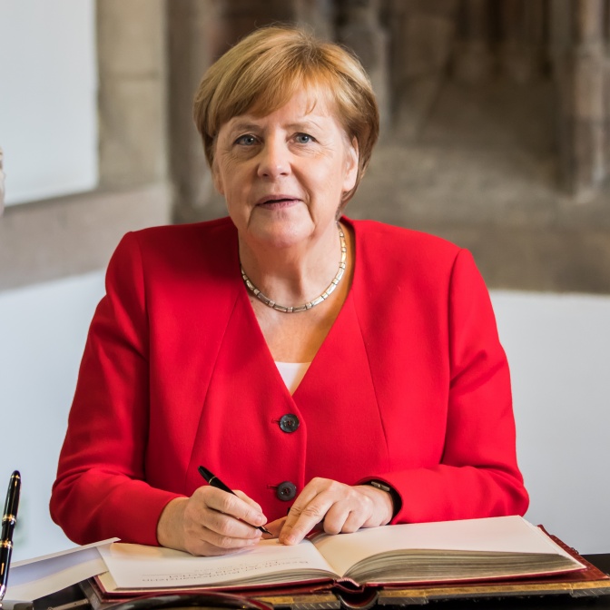 Angela Merkel sitzt vor einem großen, aufgeschlagenen Buch und hält einen Stift in der Hand.