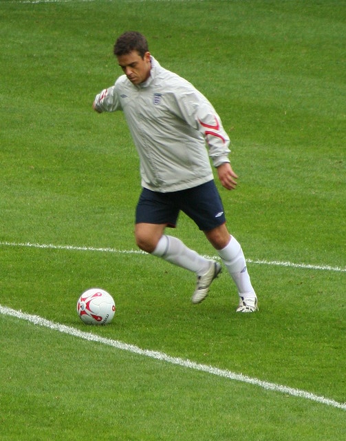 Robbie Williams in einem weißen Trikot auf dem Fußballplatz. Er spielt den Ball.