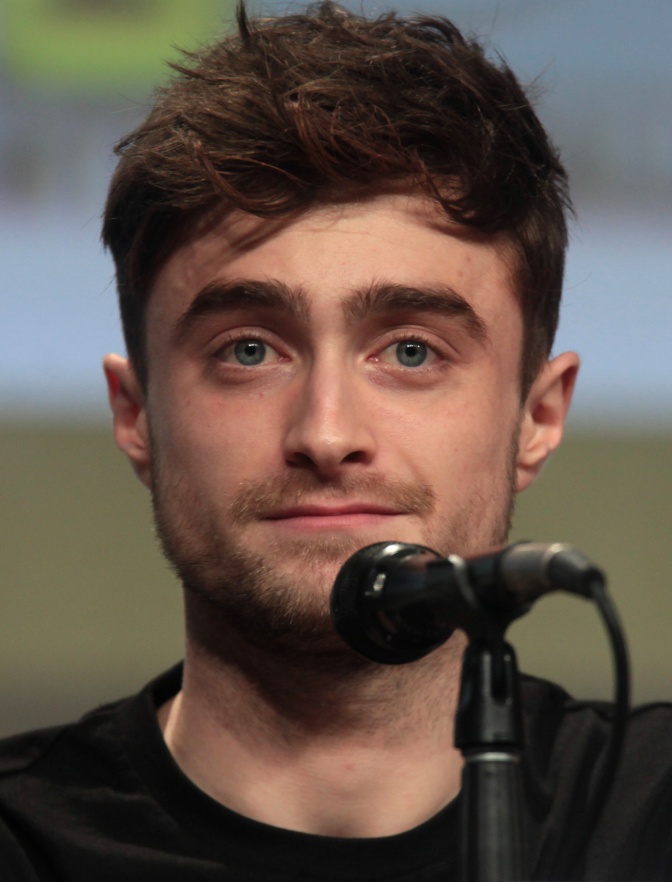 Daniel Radcliffe mit kurzen, dunklen Haaren, Er steht vor einem Mikrophon.