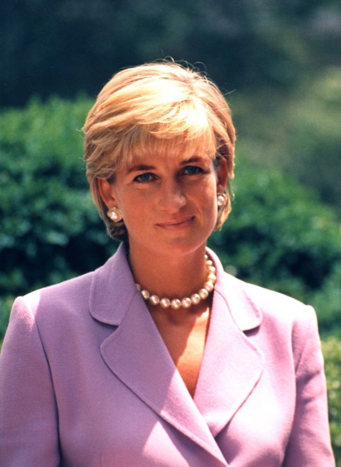Lady Diana mit blondem Kurzhaarschnitt in Kostüm und mit Perlenkette. Hinter ihr sieht man Büsche.