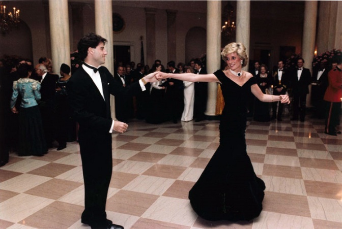 Lady Diana tanzt mit John Travolta über einen holzgetäfelten Boden. Er hält sie am ausgestreckten Arm und dreht sie. Sie trägt ein schwarzes Samtkleid.
