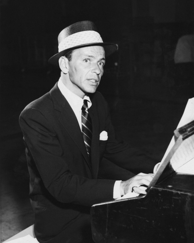 Ein schwarz-weiß-Foto von Frank Sinatra am Flügel. Er trägt Anzug, Krawatte und Hut.