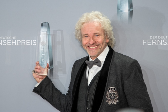 Thomas Gottschalk in Anzug und Fliege. Er steht lächelnd vor einer Logowand und hält eine Trophäe aus Glas in der Hand. Auf der Logowand steht der Schriftzug Deutscher Fernsehpreis.