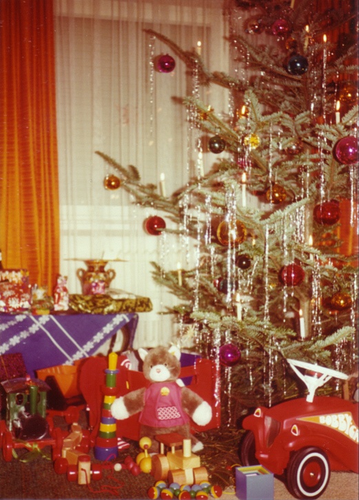 Ein geschmückter Baum steht vor einem Fenster mit Gardinen. Darunter stehen Geschenke, unter anderem ein Bobby-Car.