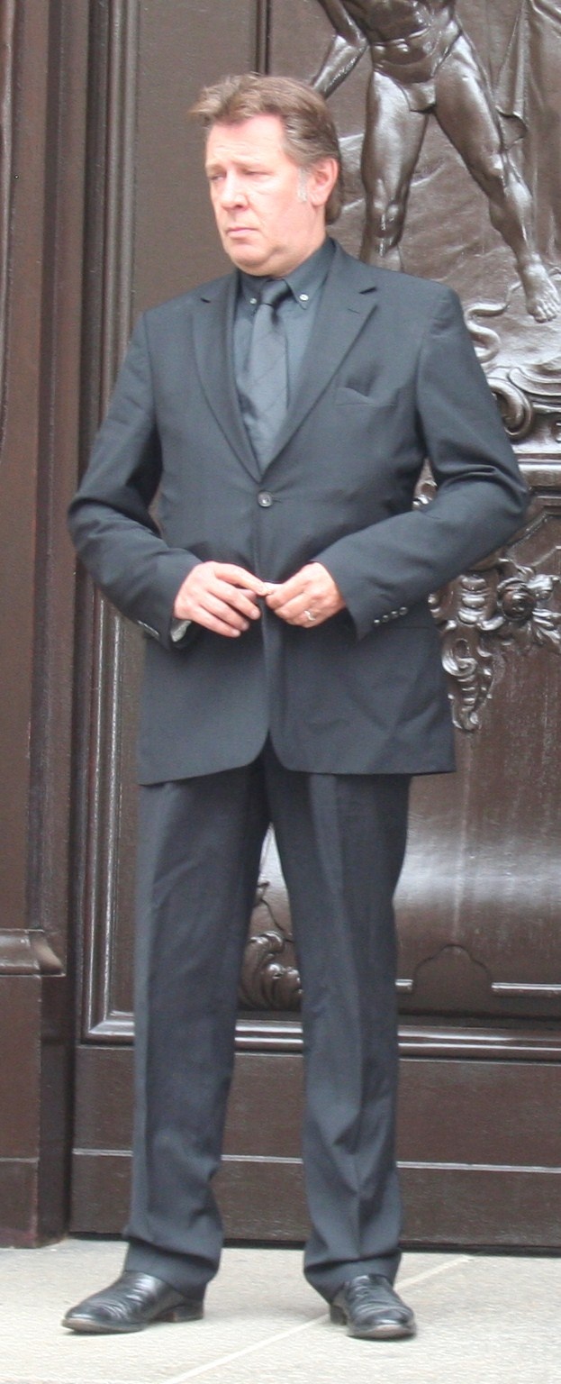 Jan Fedder steht im Anzug vor einer schweren Holztür