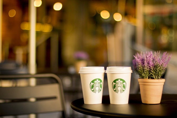 2 Pappbecher mit Starbuckslogo stehen auf einem Tisch neben einem kleinen Blumentopf