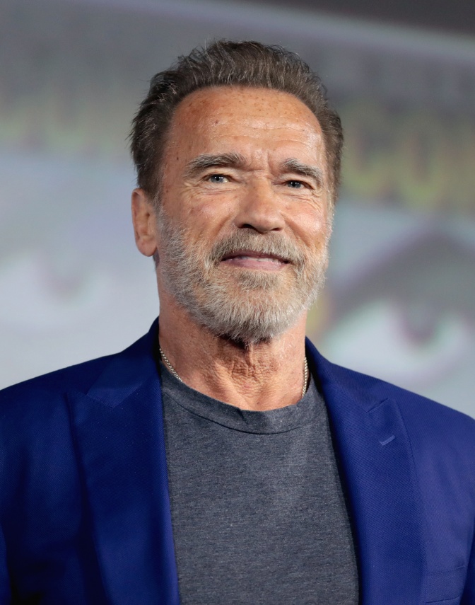 Arnold Schwarzenegger mit braunen Haaren und grauem Vollbart. Er trägt ein schlichtes Shirt und ein blaues Sakko.