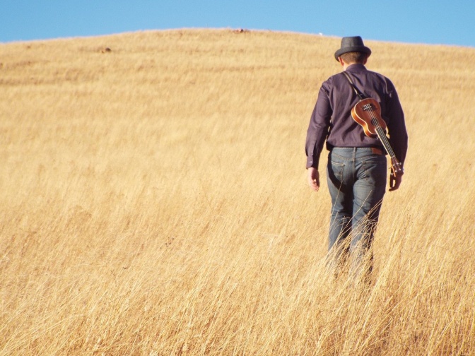 Ein Mann läuft mit einer Ukulele auf dem Rücken durch ein Kornfeld.