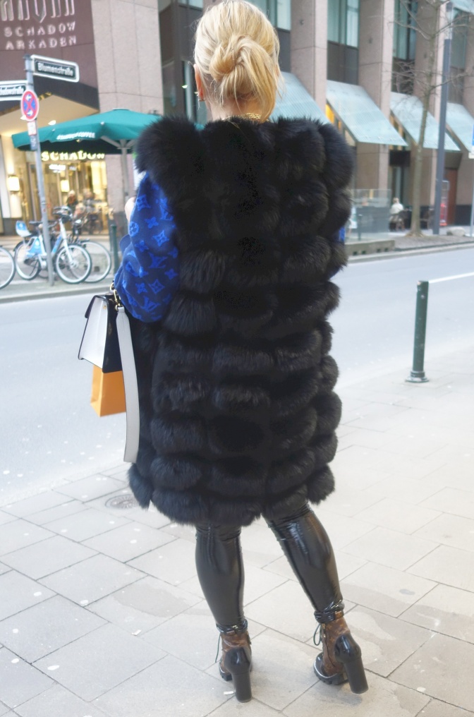 Eine von hinten fotografierte Frau in einer langen, dunklen Pelzweste. Sie steht in der Fußgängerzone einer Stadt.