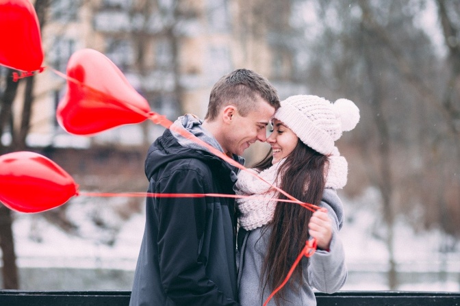 Ein Mann und eine Frau tragen Winterkleidung und lächeln sich an. Ihre Nasen berühren sich dabei. Sie hält rote Herzluftballons in der Hand.