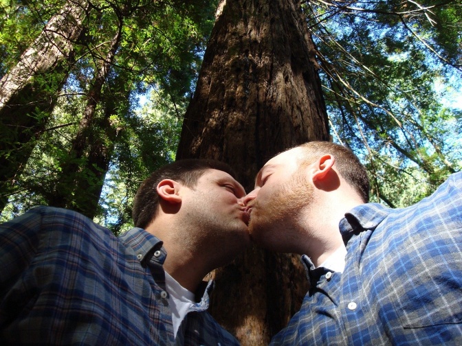 2 Männer in einheitlichen Karohemden küssen sich. Im Hintergrund sieht man einen großen Laubbaum.