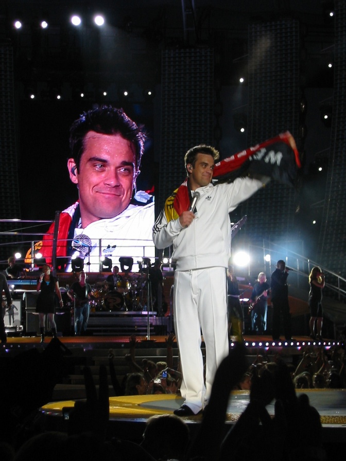 Robbie Williams steht auf der Bühne und steckt einen Schal in die Höhe. Hinter ihm sieht man einen Momitor, auf dem sein Bild groß projiziert wird.
