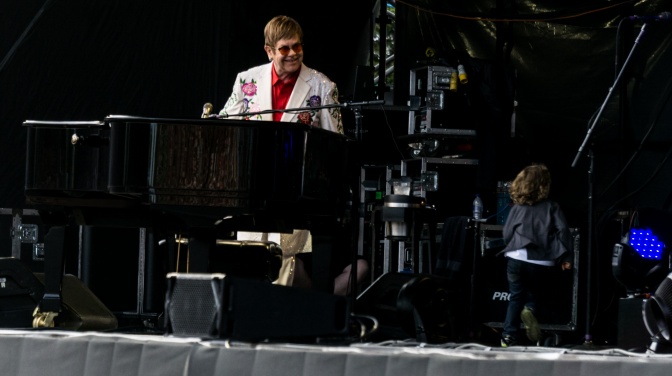 Elton John am Klavier mit farbiger Sonnenbrille und einem Sakko mit Blumenmuster.