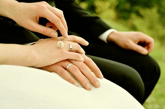 Die Hände eines Ehepaars in Hochzeitskleid und Anzug. Sie tragen beide Eheringe und haben die Hände ineinander gelegt.