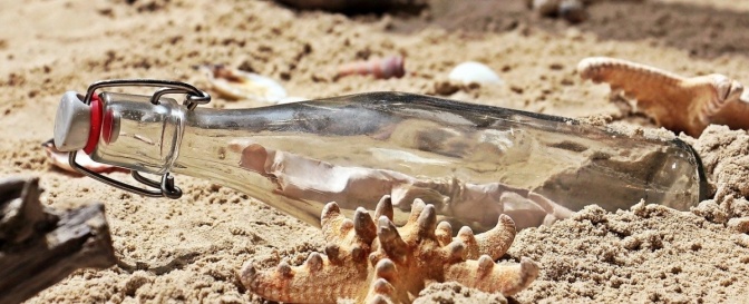 Eine Flasche mit einem Brief darin liegt im Sand, daneben liegen Muscheln und Seesterne.