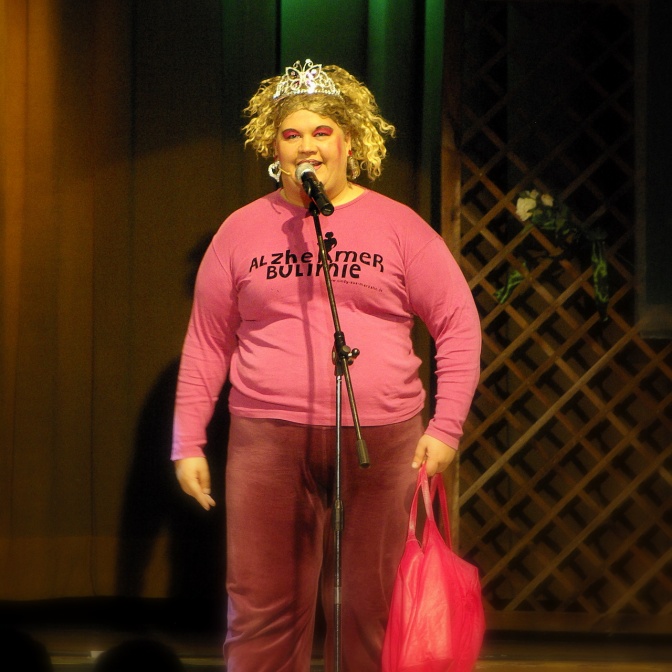Ilka Bessin in pinkfarbenem Jogginganzug und mit blonden, gewellten Haaren. Sie spricht in ein Standmikro.