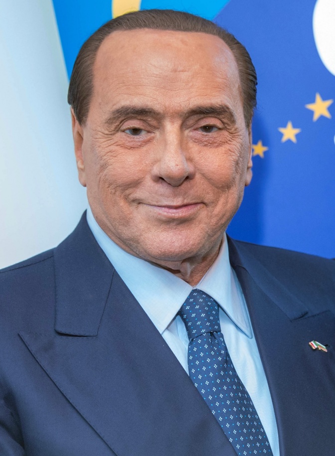 Silvio Berlusconi vor einer Europaflagge. Er hat eine Stirnglatze und trägt Anzug und Krawatte.