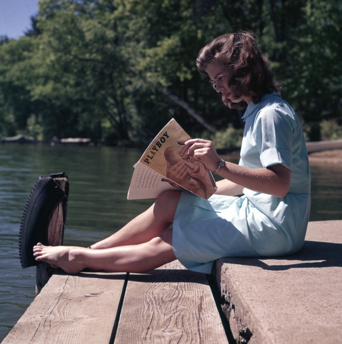 Eine Frau sitzt auf einem Steg am Wasser und liest die Zeitschrift Playboy.