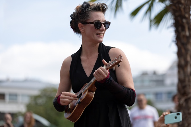 Die Musikerin Amanda Palmer spielt auf einer Ukulele. Sie hat ihre Haare hochgesteckt und trägt eine dunkle Sonnenbrille. Im Hintergrund sieht man eine Palme.