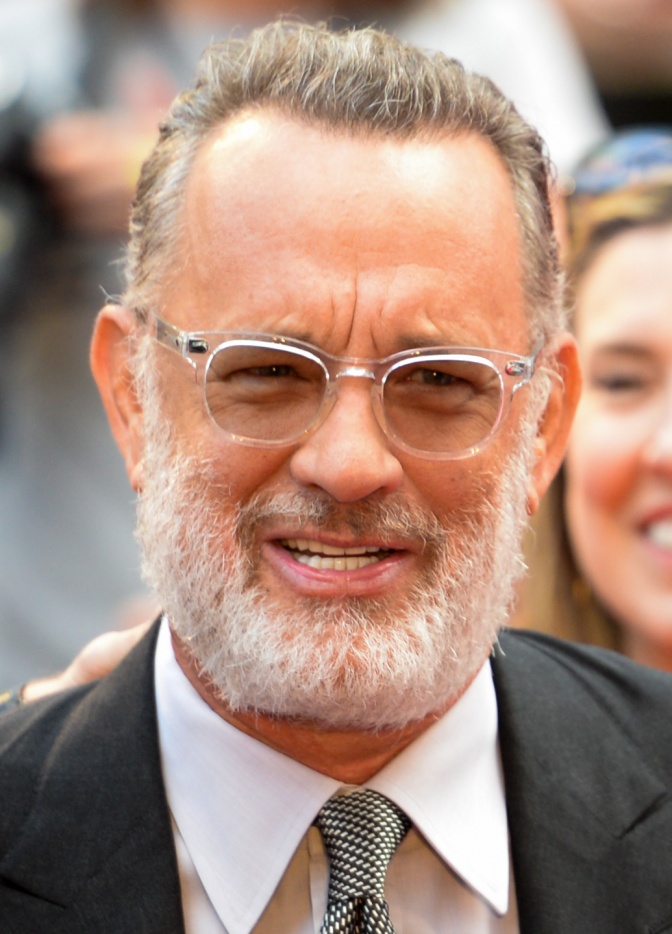 Tom Hanks mit grauem Vollbart und Brille. Er trägt Anzug und Krawatte.