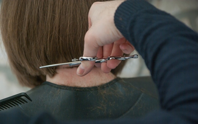 Eine Hand mit einer Schere schneidet einer Frau die Haare im Nacken auf Kinnlänge ab.