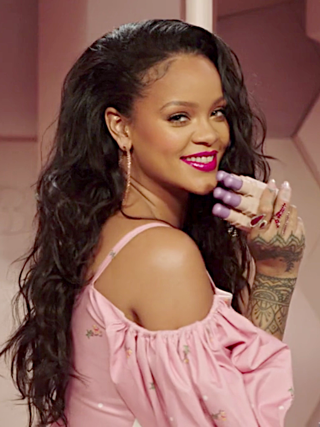 Rihanna in einer schulterfreien Bluse. Sie hat schwarze, lange gewellte Haare und lächelt. Sie hat mehrere Lippenstifte in der Hand. Ihre Hand ist mit Henna-Tattoos bemalt.
